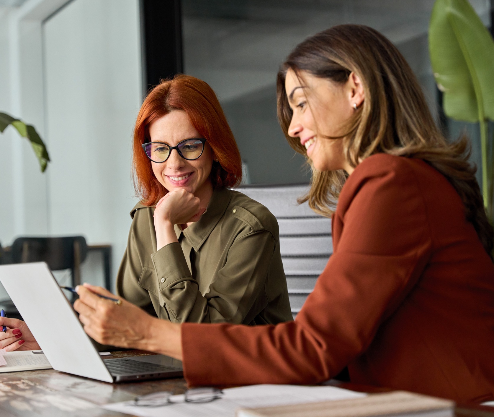 Two happy busy female employees working together using computer planning project. Middle aged professional business woman consulting teaching young employee looking at laptop sitting at desk in office