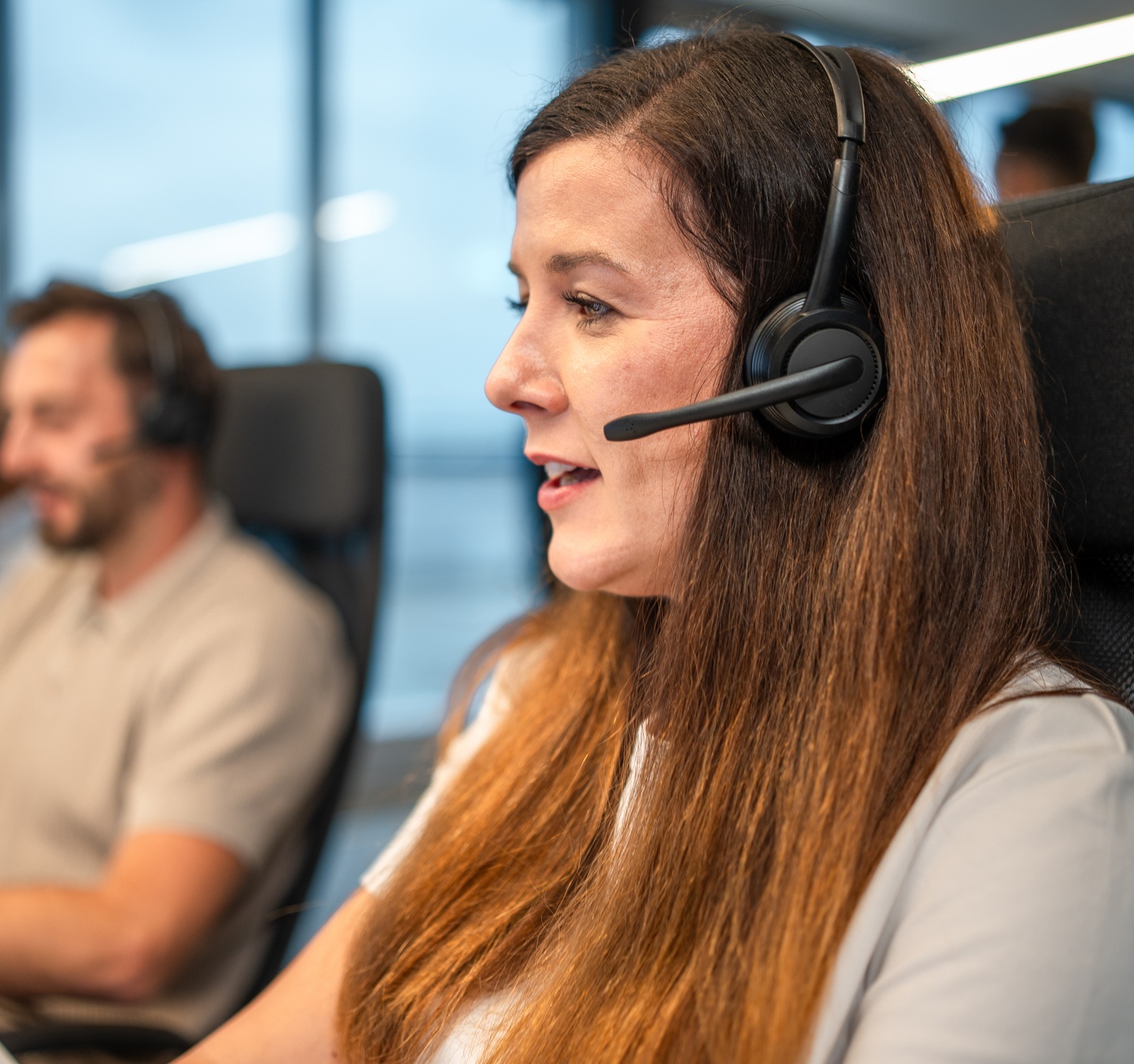 Mid adult Caucasian female professional wearing a headset and communicating in an open plan office setting, focused on managing tasks and collaborating with colleagues.